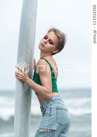Portrait of sensual beautiful woman with closed eyes holding white surfboard with both hands, posing sexy. Fashion model with short hair and bright makeup wearing casual clothing - top and blue jeans Portrait of sensual beautiful woman with closed eyes holding white surfboard with both hands, posing sexy. Fashion model with short hair and bright makeup wearing casual clothing - top and blue jeans 111918366
