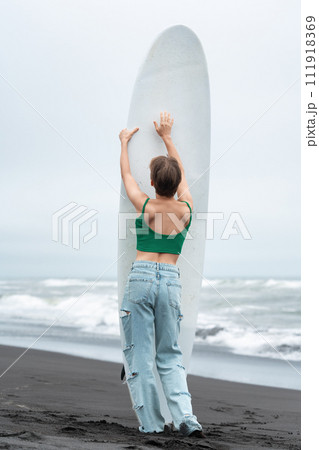 Rear view of woman surfer arms raised and holding surfboard vertically. Unrecognizable female standing on sandy beach, posing on background of ocean waves. Hipster woman wearing top and blue jeans 111918369