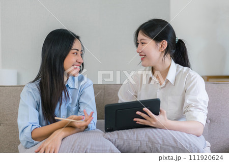 Two millennial Asian females looking at a digital tablet together in a living room. 111920624