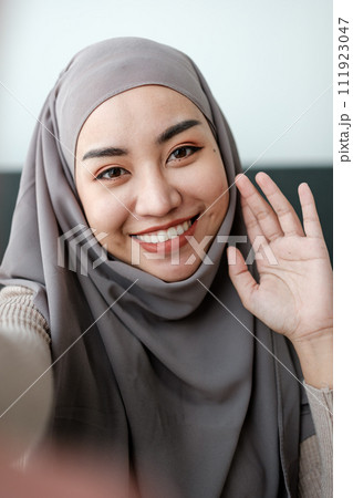 A Beautiful asian woman in hijab talks on video call, online meeting with colleagues and clients, woman cheerfully waves her hand and greets interlocutors online. 111923047