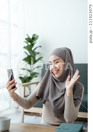 A Beautiful asian woman in hijab talks on video call, online meeting with colleagues and clients, woman cheerfully waves her hand and greets interlocutors online. 111923079