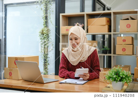 A portrait of a successful young Asian Muslim female online seller sits at her working desk with a laptop and cardboard boxes on a table. 111940208