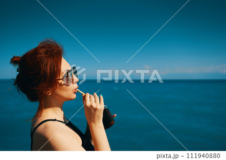 Woman on summer vacation. Red-haired girl in sunglasses drinks lemonade from bottle through cocktail tube. Blue sea, sunlight and clear sky on background. Copy space for travel agency advertising. Woman on summer vacation. Red-haired girl in sunglasses drinks lemonade from bottle through cocktail tube. Blue sea, sunlight and clear sky on background. Copy space for travel agency advertising. 111940880