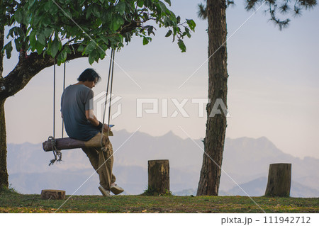 landscape and travel concept with solo freelancer man use tablet work at wooden swing under large tree and layer of mountain 111942712