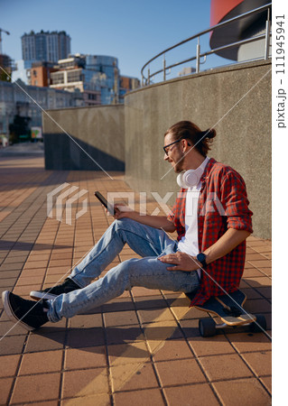 Hipster man with longboard having rest while sitting on steps and using phone Hipster man with longboard having rest while sitting on steps and using phone 111945941