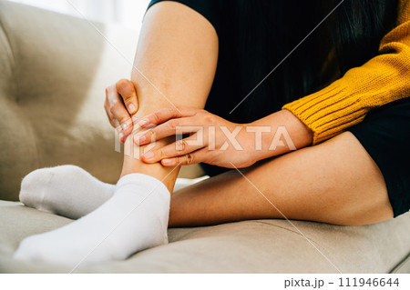 Woman seated on sofa clutching her painful ankle portraying discomfort. Reflecting health care varicose vein prevention and emphasizing leg recovery and pain relief concept. medical 111946644