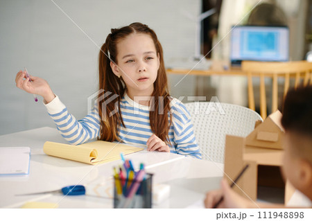 Serious schoolgirl taking to classmate and writing in copybook Serious schoolgirl taking to classmate and writing in copybook 111948898