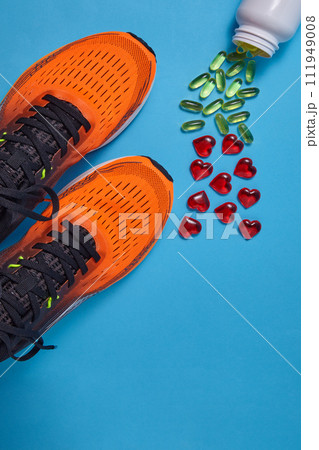 Orange sports sneakers and yellow vitamin D capsules and red glass hearts are scattered on a blue background. Vitamin supplements and physical activity to improve immunity 111949008