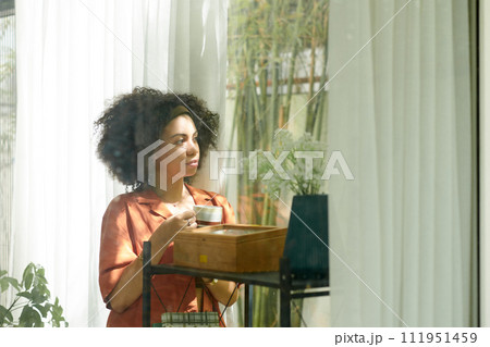 Pensive Latin woman drinking cup of coffee during break and looking outside through window 111951459