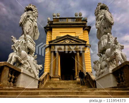 The image shows a person ascending steps towards a neoclassical building flanked by two intricate statues under a stormy sky. The image shows a person ascending steps towards a neoclassical building flanked by two intricate statues under a stormy sky. 111954373