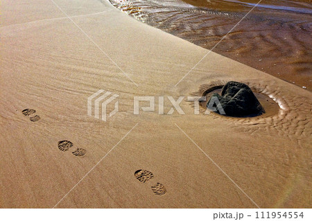A sandy beach with distinct footprints leading to a partially submerged dark rock. A sandy beach with distinct footprints leading to a partially submerged dark rock. 111954554
