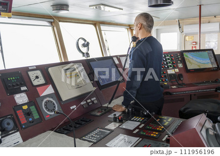 Officer on watch with radio on the navigational bridge. Caucasian man in blue uniform sweater on the bridge of cargo ship. Officer on watch with radio on the navigational bridge. Caucasian man in blue uniform sweater on the bridge of cargo ship. 111956196