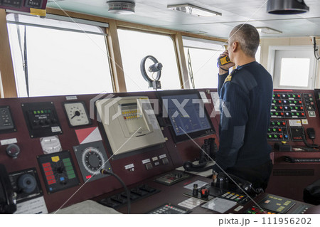 Officer on watch with radio on the navigational bridge. Caucasian man in blue uniform sweater on the bridge of cargo ship. Officer on watch with radio on the navigational bridge. Caucasian man in blue uniform sweater on the bridge of cargo ship. 111956202