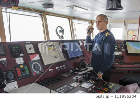 Officer on watch with radio on the navigational bridge. Caucasian man in blue uniform sweater on the bridge of cargo ship. 111956204