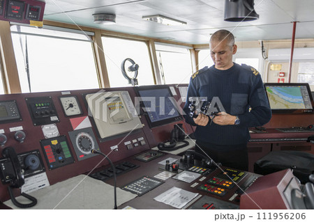 Officer on watch with binoculars on the navigational bridge. Caucasian man in blue uniform sweater on the bridge of cargo ship. Officer on watch with binoculars on the navigational bridge. Caucasian man in blue uniform sweater on the bridge of cargo ship. 111956206