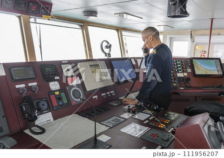 Officer on watch with radio on the navigational bridge. Caucasian man in blue uniform sweater on the bridge of cargo ship. Officer on watch with radio on the navigational bridge. Caucasian man in blue uniform sweater on the bridge of cargo ship. 111956207