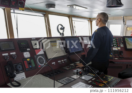 Officer on watch on the navigational bridge. Caucasian man in blue uniform sweater on the bridge of cargo ship. Officer on watch on the navigational bridge. Caucasian man in blue uniform sweater on the bridge of cargo ship. 111956212