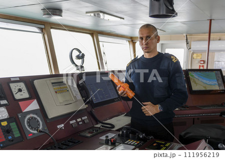 Officer on watch with SART on the navigational bridge. Caucasian man in blue uniform sweater using search and rescue radar transponder on the bridge of cargo ship. Officer on watch with SART on the navigational bridge. Caucasian man in blue uniform sweater using search and rescue radar transponder on the bridge of cargo ship. 111956219