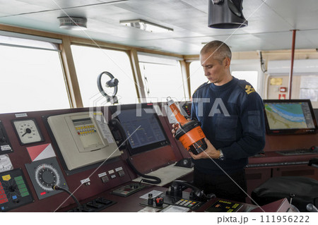 Officer on watch with EPIRB on the navigational bridge. Caucasian man in blue uniform sweater using emergency position indicating radio beacon on the bridge of cargo ship. Officer on watch with EPIRB on the navigational bridge. Caucasian man in blue uniform sweater using emergency position indicating radio beacon on the bridge of cargo ship. 111956222