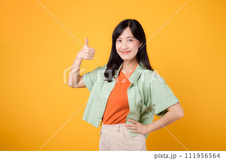 captivating young Asian woman in her 30s, dressed in an orange shirt and green jumper. Her thumbs up gesture, isolated on a sunny yellow background, embodies the concept of support. captivating young Asian woman in her 30s, dressed in an orange shirt and green jumper. Her thumbs up gesture, isolated on a sunny yellow background, embodies the concept of support. 111956564