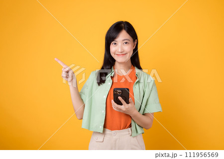 young 30s asian woman happy smile dressed in orange shirt showing smartphone with pointing finger hand gesture to free space isolated on yellow studio background. app smartphone concept young 30s asian woman happy smile dressed in orange shirt showing smartphone with pointing finger hand gesture to free space isolated on yellow studio background. app smartphone concept 111956569