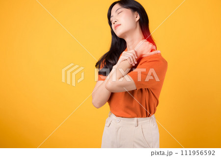 Vigilant Asian woman in her 30s, wearing an orange shirt, holds her painful neck on yellow background. office syndrome health care concept. 111956592