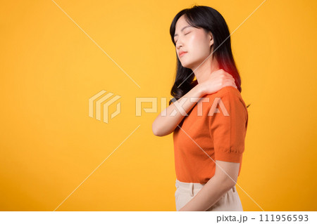 Vigilant young Asian woman in her 30s, wearing an orange shirt, holds her pain shoulder on yellow background. Neck ache therapy medical office syndrome concept. Vigilant young Asian woman in her 30s, wearing an orange shirt, holds her pain shoulder on yellow background. Neck ache therapy medical office syndrome concept. 111956593