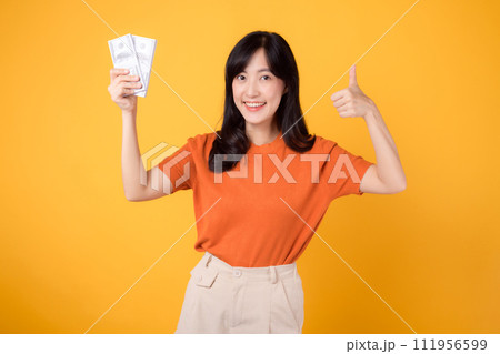Vivacious young Asian woman in her 30s, thumb up hand gesture to cash money dollars, standing on vibrant yellow backdrop. 111956599
