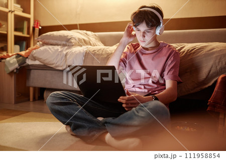 Full length portrait of young teenage boy using laptop while sitting on floor at night with legs crossed and wearing wireless headphones Full length portrait of young teenage boy using laptop while sitting on floor at night with legs crossed and wearing wireless headphones 111958854
