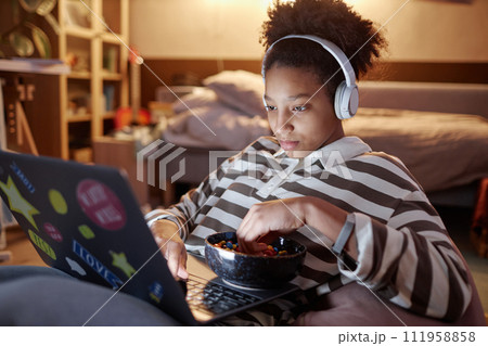 Portrait of African American teen girl using laptop at night at home and eating candy snacks 111958858