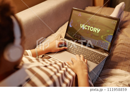 Close up of teenager playing online video games via laptop with victory screen while lying on bed at night Close up of teenager playing online video games via laptop with victory screen while lying on bed at night 111958859