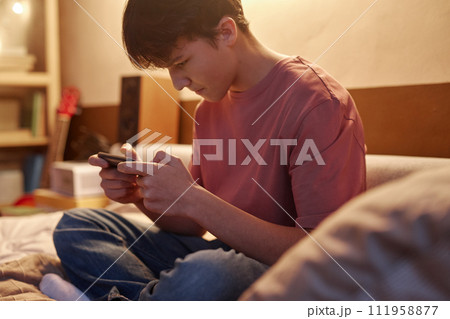 Side view portrait of young teenage boy playing mobile games at night sitting on bed in dim light Side view portrait of young teenage boy playing mobile games at night sitting on bed in dim light 111958877