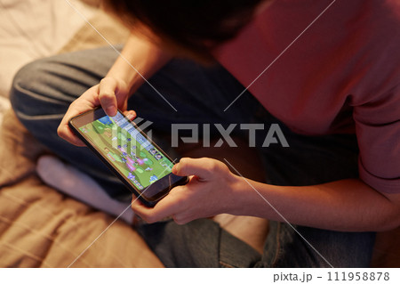 Close up of unrecognizable teenage boy playing mobile game online via smartphone sitting on bed at night Close up of unrecognizable teenage boy playing mobile game online via smartphone sitting on bed at night 111958878