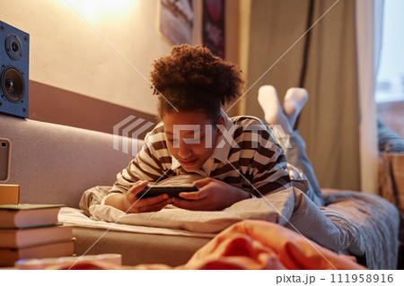 Portrait of African American teenage girl playing mobile game lying on bed at home and smiling 111958916