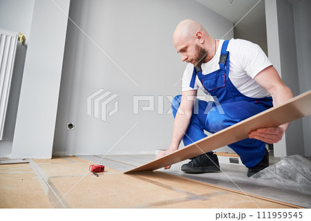 Male construction worker laying laminate wooden plank on floor underlayment in apartment under renovation. Man in workwear installing laminate timber flooring in living room. Home renovation concept. Male construction worker laying laminate wooden plank on floor underlayment in apartment under renovation. Man in workwear installing laminate timber flooring in living room. Home renovation concept. 111959545