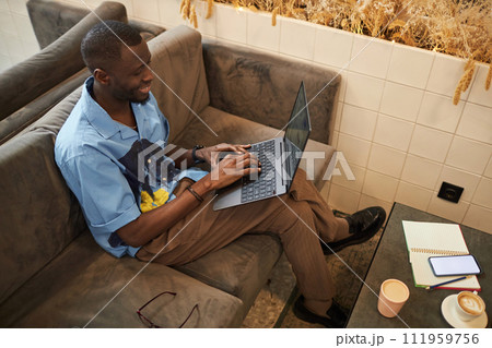 High angle portrait of smiling Black man using laptop sitting on couch in office lounge and networking online, copy space 111959756