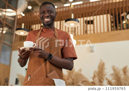 Waist up portrait of smiling Black man as barista holding coffee cup in cafe and wearing apron, copy space Waist up portrait of smiling Black man as barista holding coffee cup in cafe and wearing apron, copy space 111959765