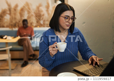 Portrait of Middle Eastern young woman wearing glasses using laptop in coffee shop and holding cup of coffee while working online copy space 111959817