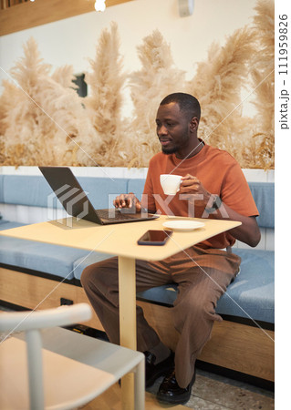 Vertical full length portrait of Black young man using laptop at table in coffee shop holding cup of coffee Vertical full length portrait of Black young man using laptop at table in coffee shop holding cup of coffee 111959826
