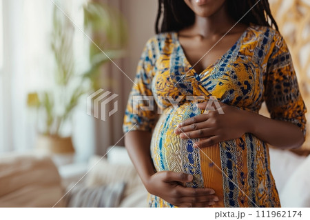 9 month pregnant African American woman wears colorful dress touching her belly at home 111962174