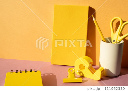 Office supply stationery. Yellow notebook, colored pencil, scissors, tape dispenser, clip on pink desk. orange wall background. workspace 111962330