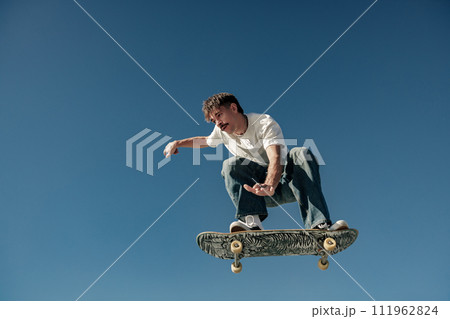 Active man doing tricks in the air on his skateboard at the skate park on blue sky background 111962824