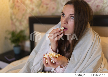 Young woman watching movie on TV in the bed and eating popcorn Young woman watching movie on TV in the bed and eating popcorn 111963443