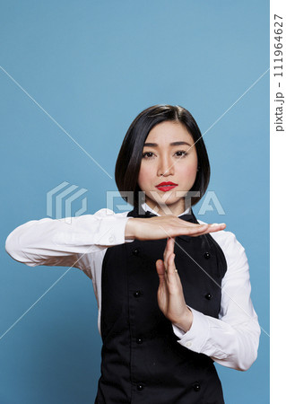 Serious asian waitress in uniform showing timeout sign with hands, asking to take break portrait. Young woman receptionist making interruption and pause gesture while looking at camera Serious asian waitress in uniform showing timeout sign with hands, asking to take break portrait. Young woman receptionist making interruption and pause gesture while looking at camera 111964627