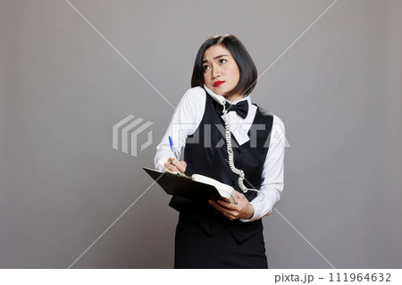 Catering service employee frowning while speaking on landline phone with client and taking notes on clipboard. Waitress looking upwards while chatting with cafe manager on telephone 111964632