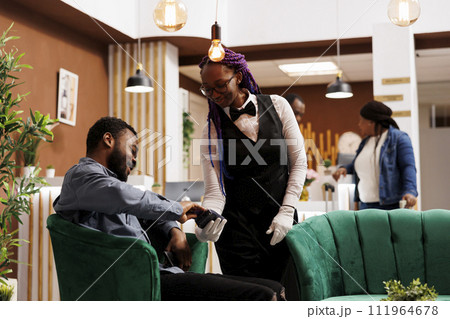 Smiling African American man tourist holding credit card paying bill while resting relaxing in hotel lounge area. Friendly female waitress holding pos terminal serving guest at hotel lobby 111964678