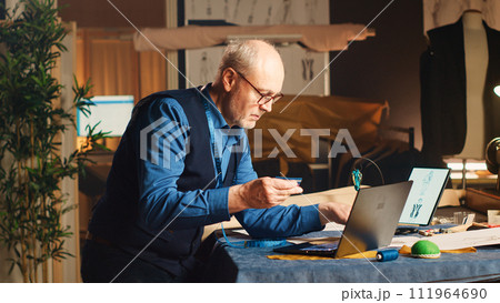 Fashion designer buying fabric online with credit card at laptop, doing online shopping to purchase material and sewing tools for custom made creation. Professional tailor in atelier. Handheld shot. 111964690