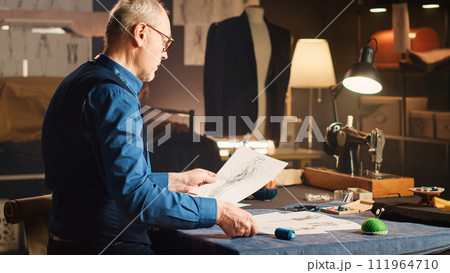 Senior tailor examining sketches with items and sewing tools, working on making clothes in luxury atelier. Professional old couturier using drawings and fabric to create designs. 111964710