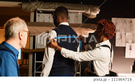Tailors team taking measurements for client, working in atelier to create custom made tailored suit. Couturier and assistant measuring customer to design perfect fit clothes. Handheld shot. 111964719
