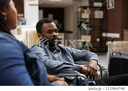 African American man tourist with physical impairment in wheelchair waiting with luggage in hotel lobby, arriving at rehabilitation center for disabled. Inclusive accessible tourism African American man tourist with physical impairment in wheelchair waiting with luggage in hotel lobby, arriving at rehabilitation center for disabled. Inclusive accessible tourism 111964746